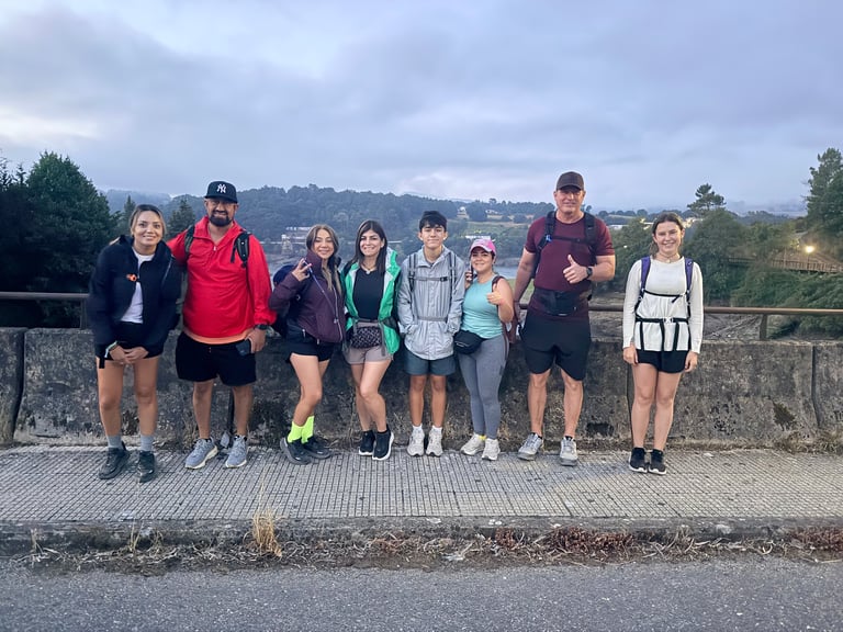 Group of eight hikers standing together on a stone wall overlooking a rural hillside landscape under cloudy skies